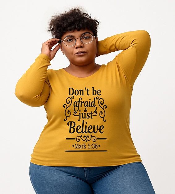 Mustard Christian long sleeve t-shirt with “Don’t Be Afraid Just Believe” Mark 5:36 scripture design, worn by woman in glasses and ripped jeans.