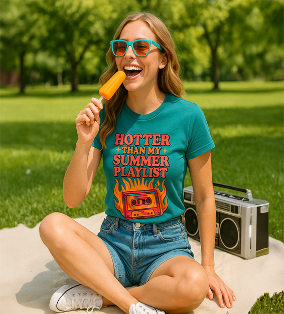 Woman wearing a teal t-shirt with 'Hotter Than My Summer Playlist' design, eating a popsicle in a park.