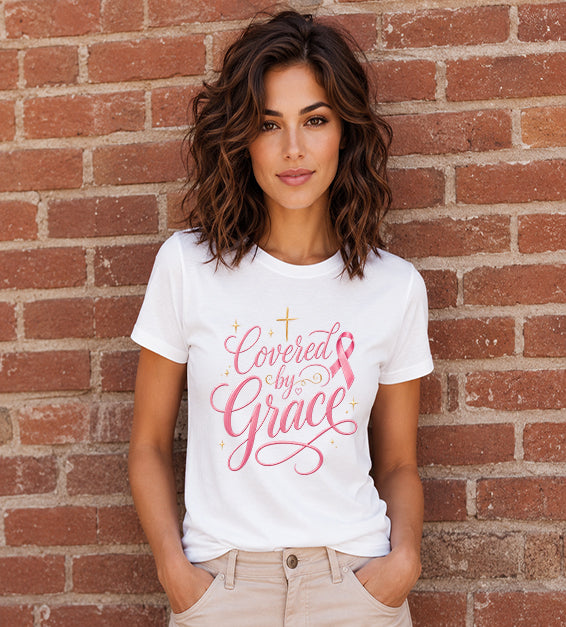 Woman wearing a white Bella + Canvas 3001 t-shirt featuring the "Covered by Grace" pink ribbon Christian graphic with an elegant gold cross standing in front of a red brick wall.