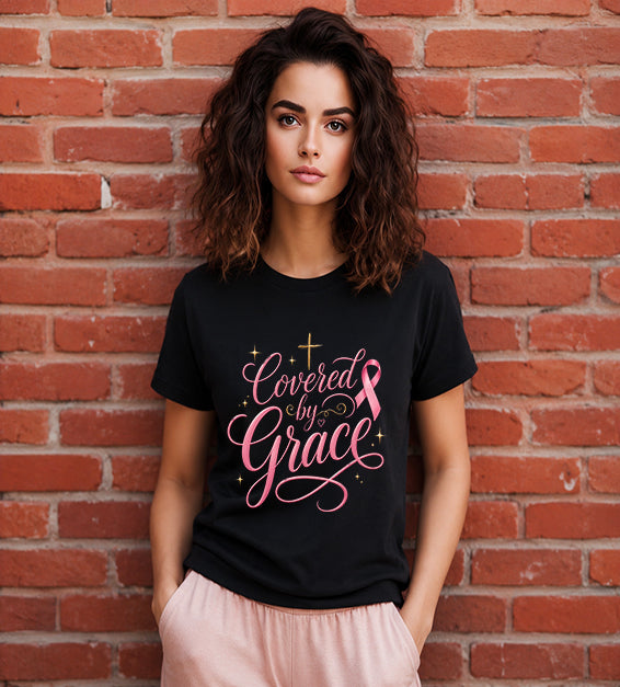 Woman wearing a black Bella + Canvas 3001 t-shirt featuring the "Covered by Grace" pink ribbon Christian graphic with an elegant gold cross standing in front of a red brick wall.