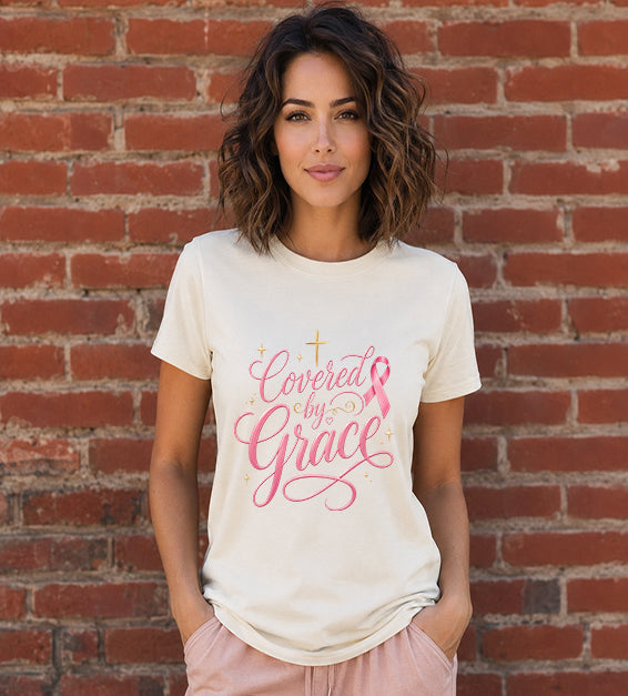 Woman wearing a natural Bella + Canvas 3001 t-shirt featuring the "Covered by Grace" pink ribbon Christian graphic with an elegant gold cross standing in front of a red brick wall.