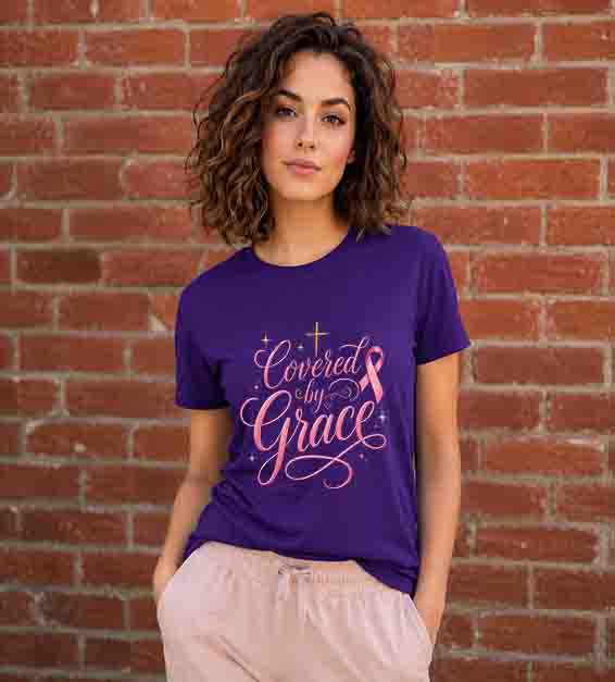Woman wearing a purple Bella + Canvas 3001 t-shirt featuring the "Covered by Grace" pink ribbon Christian graphic with an elegant gold cross standing in front of a red brick wall.
