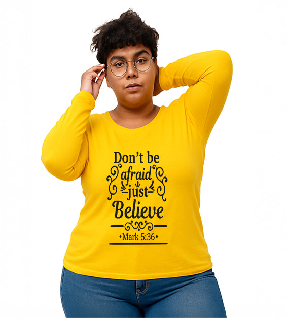 Gold Christian long sleeve t-shirt with “Don’t Be Afraid Just Believe” Mark 5:36 scripture design, worn by woman in glasses and ripped jeans.

