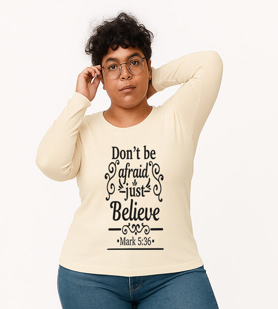 Soft Cream Christian long sleeve t-shirt with “Don’t Be Afraid Just Believe” Mark 5:36 scripture design, worn by woman in glasses and ripped jeans.
