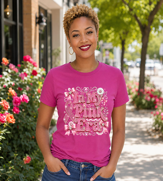 Smiling woman wearing a berry/magenta graphic t-shirt featuring the phrase "In My Pink Era" with elegant pink ribbon artwork, bows, rhinestone-inspired lettering, and feminine decorative accents while standing outdoors.