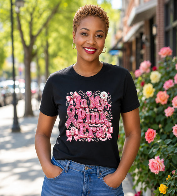 Smiling woman wearing a black graphic t-shirt featuring the phrase "In My Pink Era" with elegant pink ribbon artwork, bows, rhinestone-inspired lettering, and feminine decorative accents while standing outdoors.
