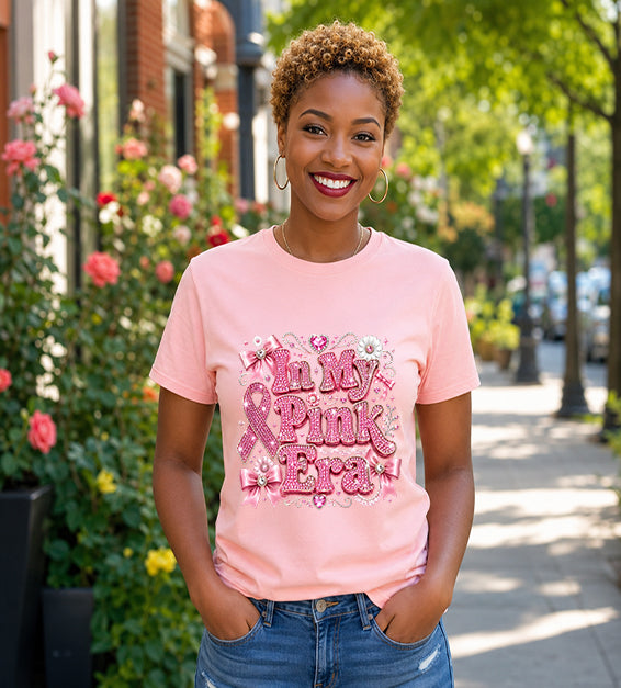 Smiling woman wearing a soft pink graphic t-shirt featuring the phrase "In My Pink Era" with elegant pink ribbon artwork, bows, rhinestone-inspired lettering, and feminine decorative accents while standing outdoors.