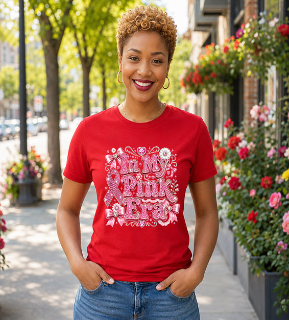 Smiling woman wearing a red graphic t-shirt featuring the phrase "In My Pink Era" with elegant pink ribbon artwork, bows, rhinestone-inspired lettering, and feminine decorative accents while standing outdoors.