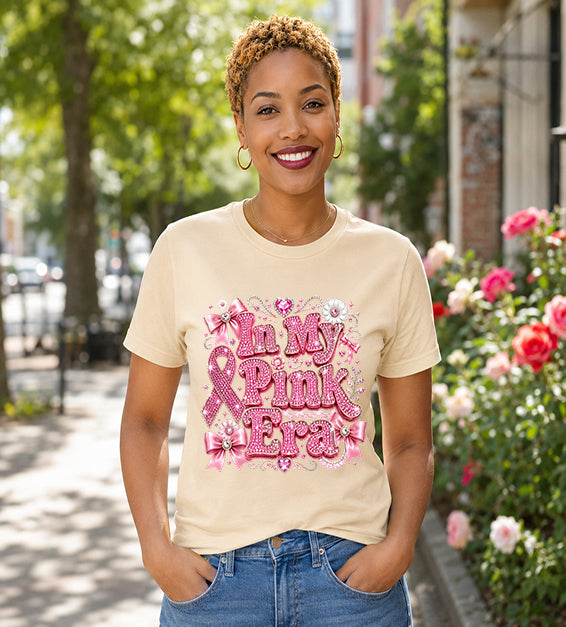 Smiling woman wearing a soft cream graphic t-shirt featuring the phrase "In My Pink Era" with elegant pink ribbon artwork, bows, rhinestone-inspired lettering, and feminine decorative accents while standing outdoors.