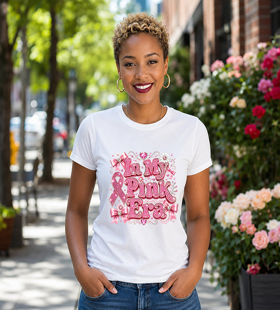 Smiling woman wearing a white graphic t-shirt featuring the phrase "In My Pink Era" with elegant pink ribbon artwork, bows, rhinestone-inspired lettering, and feminine decorative accents while standing outdoors.