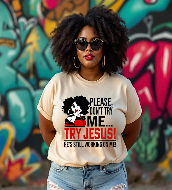 Confident  woman wearing a soft cream color T-shirt that says “Please Don’t Try Me… Try Jesus! He’s Still Working On Me” in bold black and red text, standing in front of a colorful graffiti wall.