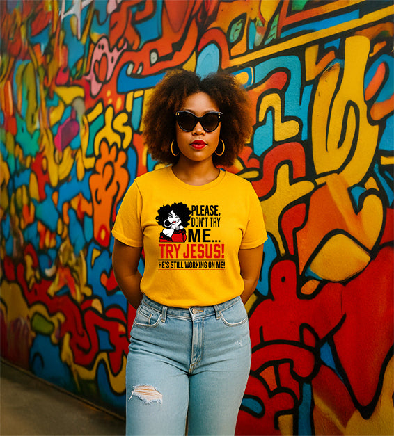 Confident woman wearing a gold/sunflower yellow color T-shirt that says “Please Don’t Try Me… Try Jesus! He’s Still Working On Me” in bold black and red text, standing in front of a colorful graffiti wall.