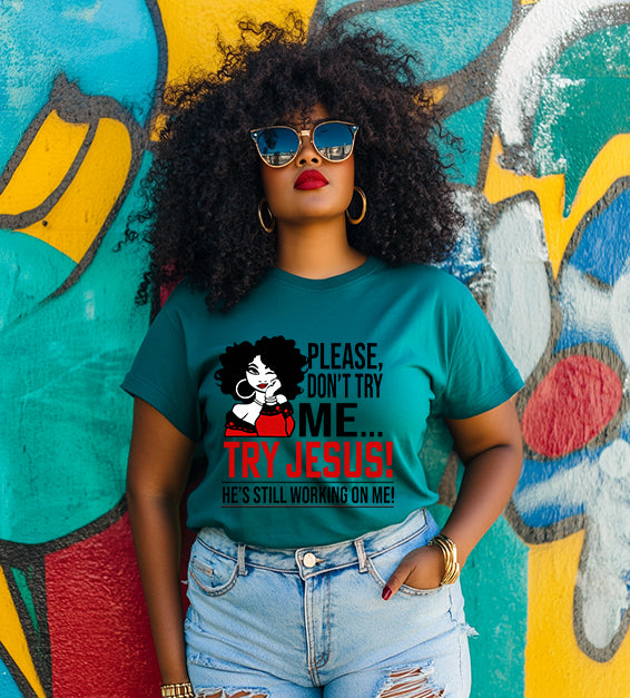 Confident woman wearing a teal color T-shirt that says “Please Don’t Try Me… Try Jesus! He’s Still Working On Me” in bold black and red text, standing in front of a colorful graffiti wall.