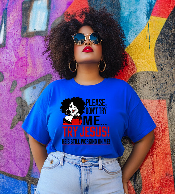 Confident woman wearing a blue/true royal color T-shirt that says “Please Don’t Try Me… Try Jesus! He’s Still Working On Me” in bold black and red text, standing in front of a colorful graffiti wall.