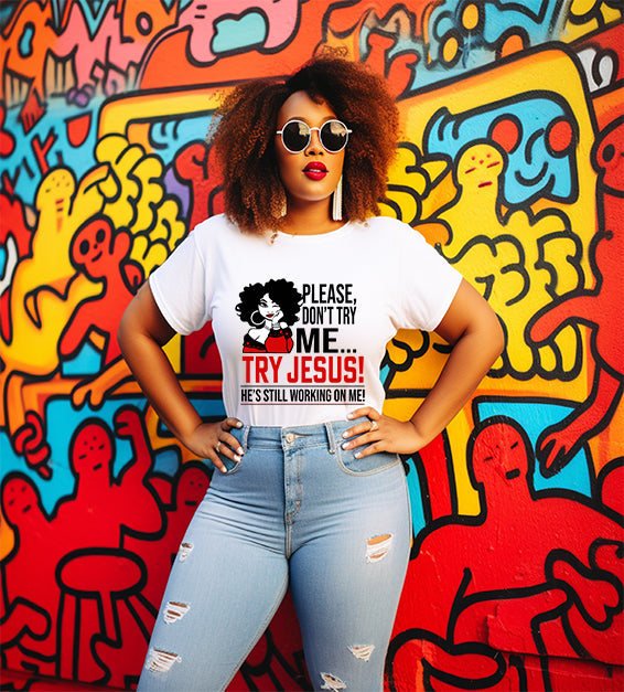 Confident woman wearing a white T-shirt that says “Please Don’t Try Me… Try Jesus! He’s Still Working On Me” in bold black and red text, standing in front of a colorful graffiti wall.
