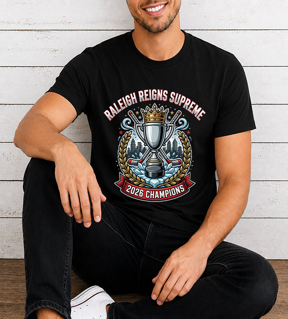 Young man wearing a black hockey championship fan t-shirt featuring a championship trophy, crown, Raleigh skyline, hockey sticks, and 2026 Champions artwork against a rustic white wooden background.