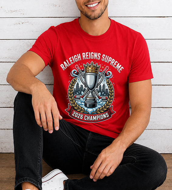 Young man wearing a red hockey championship fan t-shirt featuring a championship trophy, crown, Raleigh skyline, hockey sticks, and 2026 Champions artwork against a rustic white wooden background.