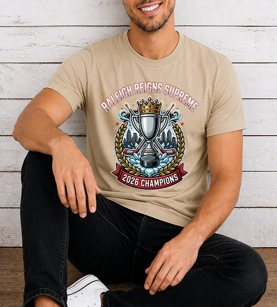 Young man wearing a sand dune hockey championship fan t-shirt featuring a championship trophy, crown, Raleigh skyline, hockey sticks, and 2026 Champions artwork against a rustic white wooden background.