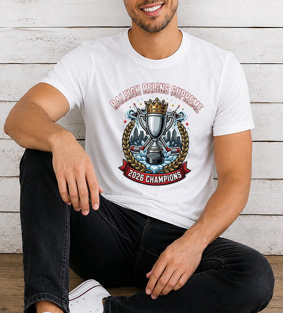 Young man wearing a white hockey championship fan t-shirt featuring a championship trophy, crown, Raleigh skyline, hockey sticks, and 2026 Champions artwork against a rustic white wooden background.