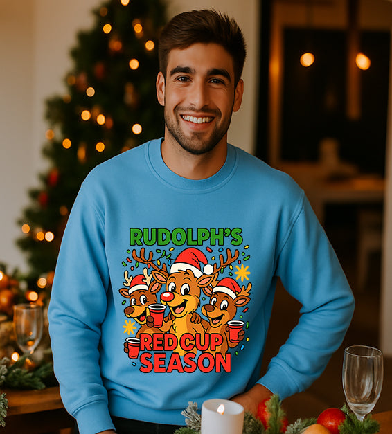 Man wearing a carolina blue Christmas sweatshirt with a festive cartoon reindeer design and the text “Rudolph’s Red Cup Season” in bright holiday colors.