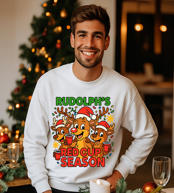 Man wearing a white Christmas sweatshirt with a festive cartoon reindeer design and the text “Rudolph’s Red Cup Season” in bright holiday colors.