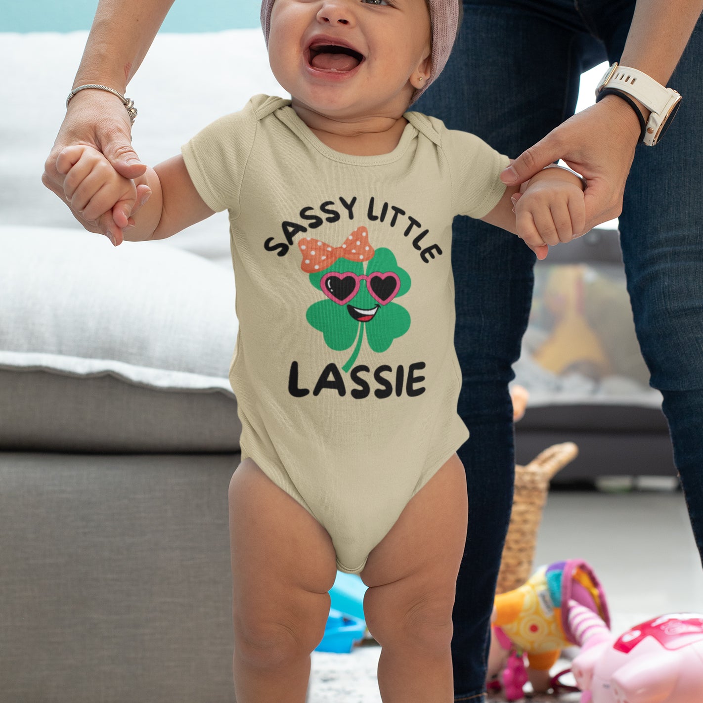 A playful green shamrock with heart-shaped pink sunglasses, a smiling face, and an orange polka-dot bow, designed for a St. Patrick’s Day infant onesie with the phrase "Sassy Little Lassie."