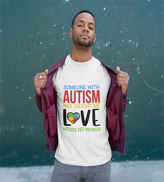 A man wearing a white T-shirt that says “Someone With Autism Has Taught Me Love Needs No Words,” featuring colorful text and a puzzle heart graphic.