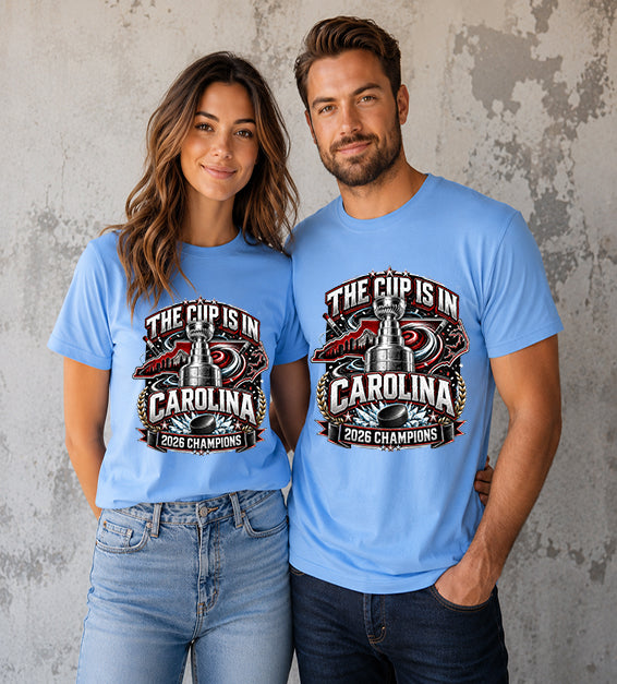 Carolina Blue t-shirt featuring The Cup Is In Carolina 2026 Champions hockey fan graphic with trophy, hockey puck, state outline, and championship artwork.
