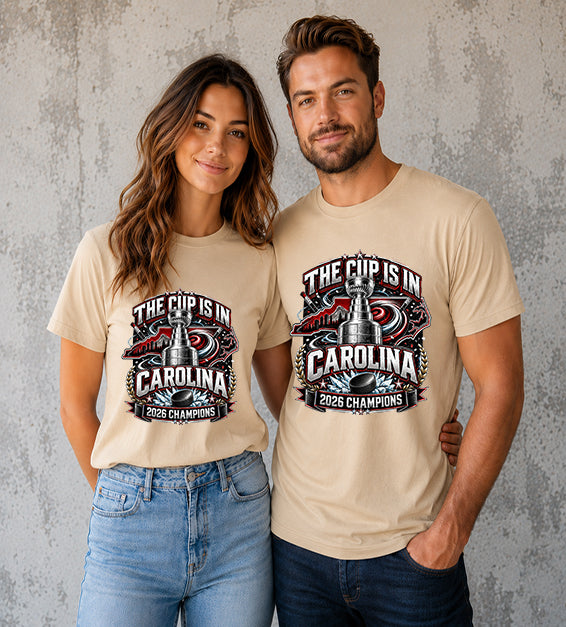 Sand Dune t-shirt featuring The Cup Is In Carolina 2026 Champions hockey fan graphic with trophy, hockey puck, state outline, and championship artwork.