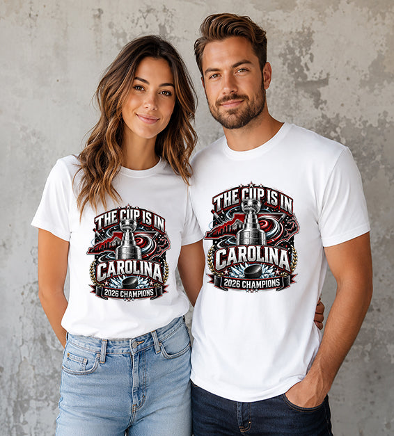 White t-shirt featuring The Cup Is In Carolina 2026 Champions hockey fan graphic with trophy, hockey puck, state outline, and championship artwork.